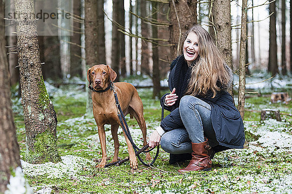 Lächelnde junge schöne Frau mit langen braunen Haaren hockt neben einem Hund an einem Baum im Wald