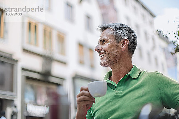 Lächelnder reifer Mann  der eine Kaffeetasse hält und wegschaut  während er in einem Straßencafé sitzt