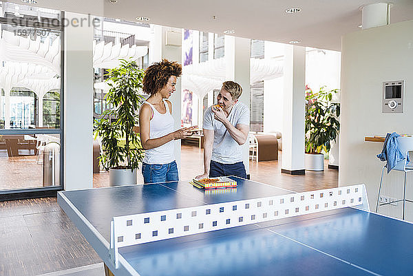 Junge Geschäftsleute essen gemeinsam Pizza zum Mittagessen in einem modernen Büro mit Tischtennisplatte