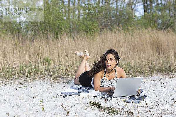 Porträt einer jungen Frau  die auf einer Decke am Strand liegt und einen Laptop benutzt