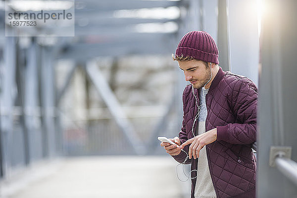 Junger Mann auf einer Brücke mit Ohrstöpseln und Smartphone