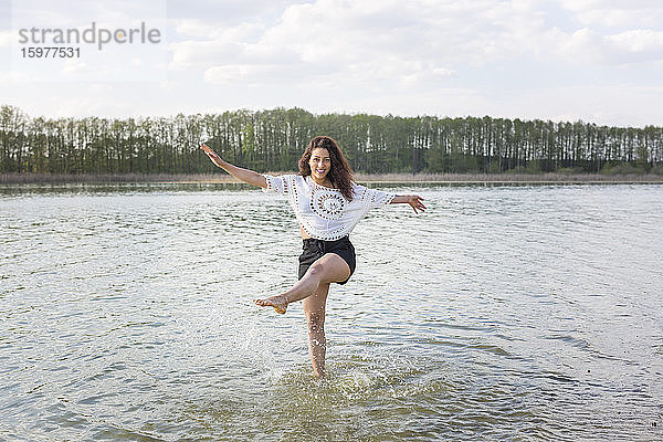 Lächelnde junge Frau steht auf einem Bein im See und spritzt mit Wasser