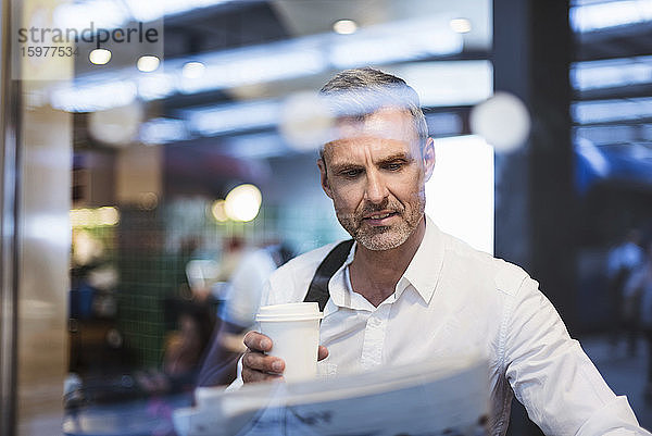 Geschäftsmann hält Kaffee und liest Zeitung  während er in einem Zug sitzt  gesehen durch ein Fenster