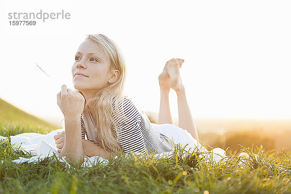 Nachdenkliche junge Frau liegt auf einer Decke gegen den klaren Himmel im Park bei Sonnenuntergang