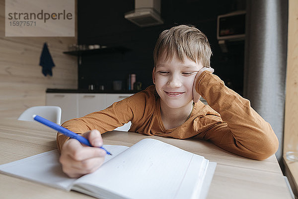 Lächelnder Junge sitzt am Tisch und schreibt zu Hause in ein Heft