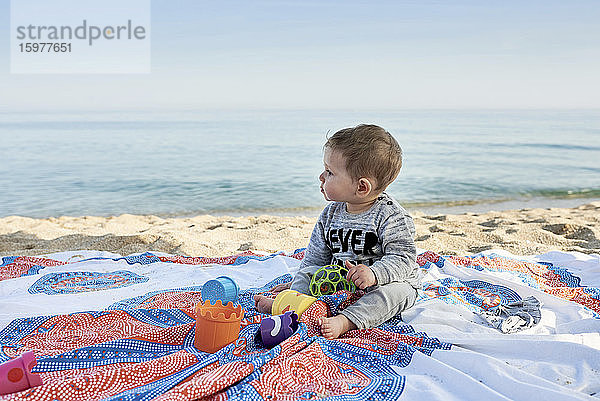 Niedlicher Junge mit Spielzeug auf einer Decke sitzend am Strand gegen das Meer