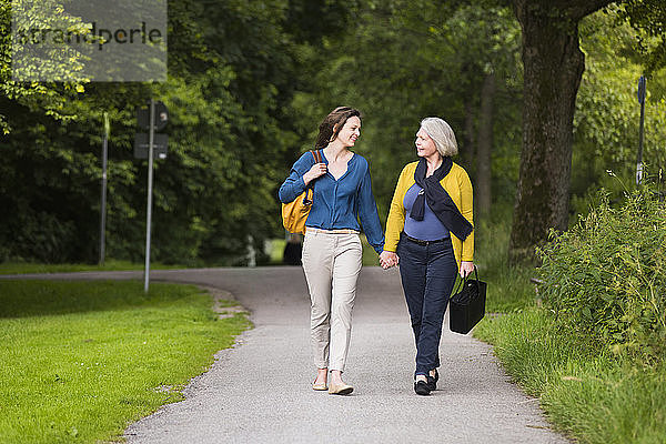 Lächelnde ältere Frau und erwachsene Tochter gehen Hand in Hand in einem Park und schauen sich an