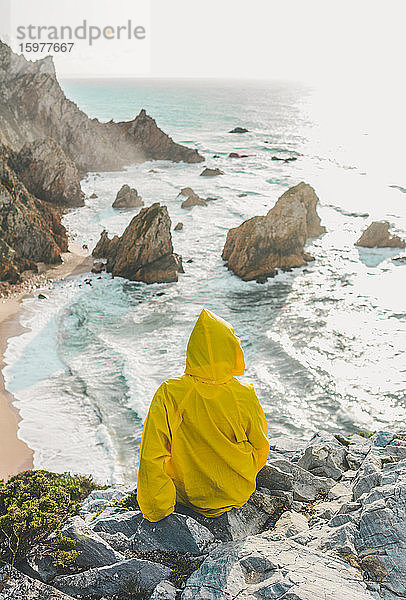 Rückansicht einer Frau  die einen Regenmantel trägt  während sie auf einem Felsen sitzt und auf den Strand schaut  Praia da Ursa  Lisboa  Portugal