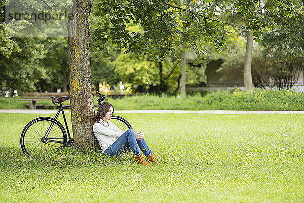 Nachdenkliche junge Frau entspannt sich  während sie mit dem Fahrrad auf einer Wiese im Park gegen einen Baum sitzt