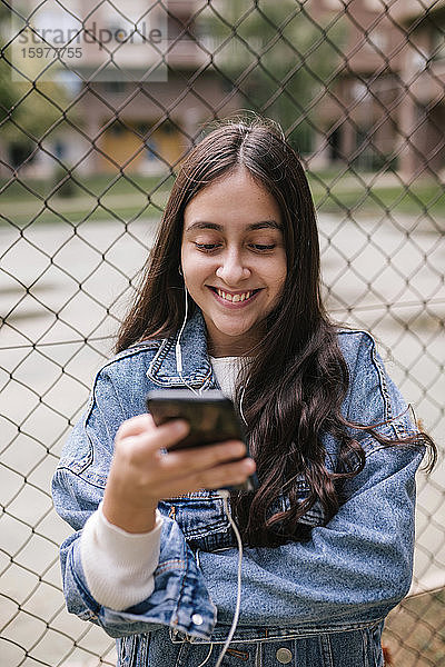Lächelndes Teenager-Mädchen  das ein Mobiltelefon benutzt  während es an einem Maschendrahtzaun auf einem Sportplatz steht