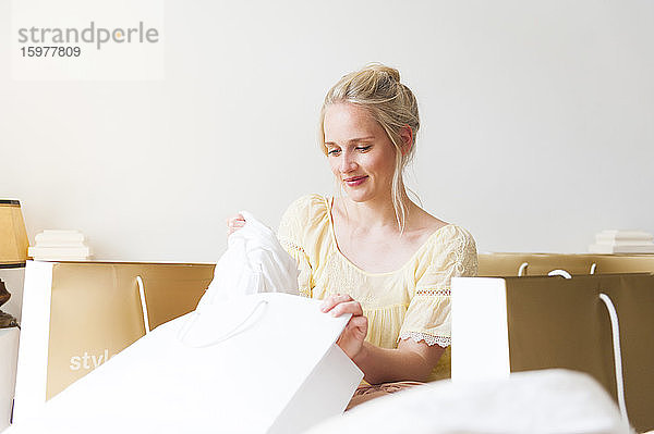 Porträt einer glücklichen blonden Frau  die mit Einkaufstüten auf dem Bett sitzt