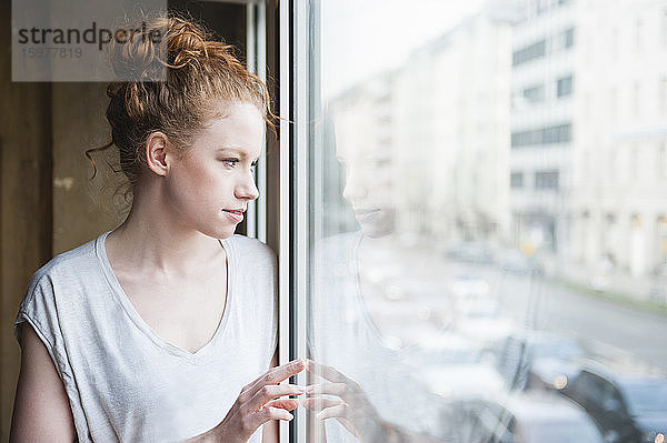 Nachdenkliche junge Frau  die durch das Fenster ihres Hauses schaut