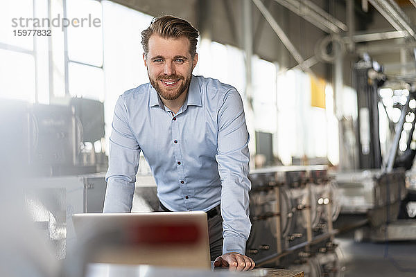 Porträt eines lächelnden jungen Geschäftsmannes mit Laptop in einer Fabrik