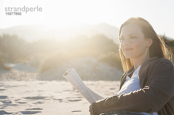 Porträt einer lächelnden Frau mit Zeitschrift  die am Strand liegt und in die Ferne schaut  Sardinien  Italien