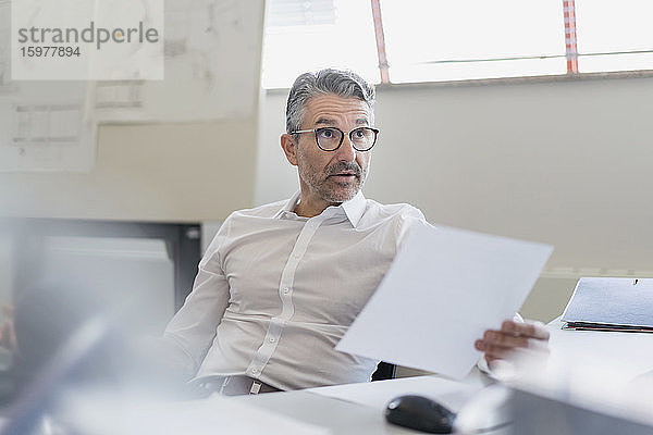 Selbstbewusster Geschäftsmann  der mit hochgezogenen Augenbrauen wegschaut  während er mit einem Dokument am Schreibtisch im Büro sitzt
