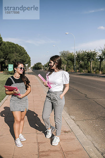 In voller Länge von lächelnden jungen Studentinnen  die Akten halten  während sie auf dem Fußweg auf dem Universitätscampus gegen den Himmel laufen