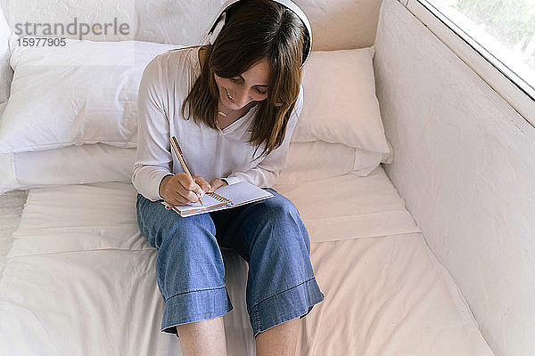 Lächelnde Frau sitzt auf dem Bett und hört Musik mit Kopfhörern  während sie Notizen macht