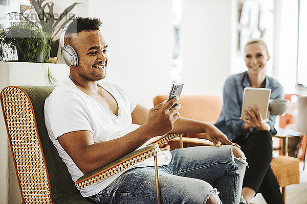 Lächelnder Mann sitzt im Sessel und hört mit Kopfhörern und Smartphone Musik