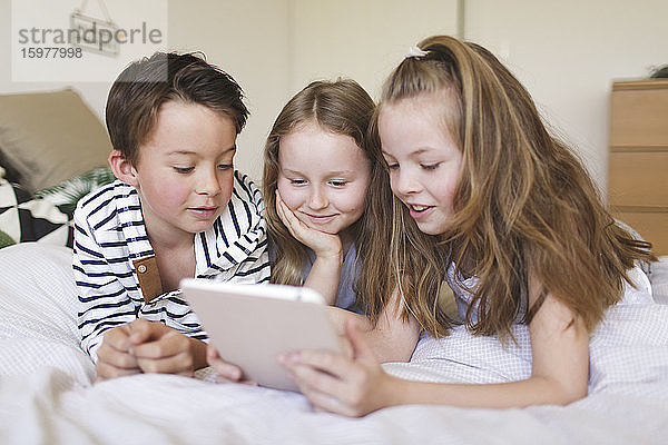 Porträt eines Jungen und seiner beiden Schwestern  die auf einem Bett liegen und ein digitales Tablet für den Hausunterricht benutzen