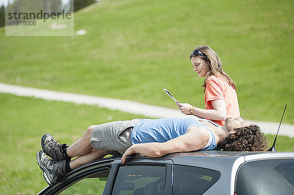 Frau liest Landkarte  während Mann auf dem Autodach bei Sonnenschein liegt