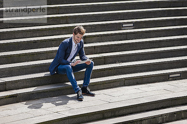 Junger Geschäftsmann sitzt auf einer Treppe und benutzt ein Tablet