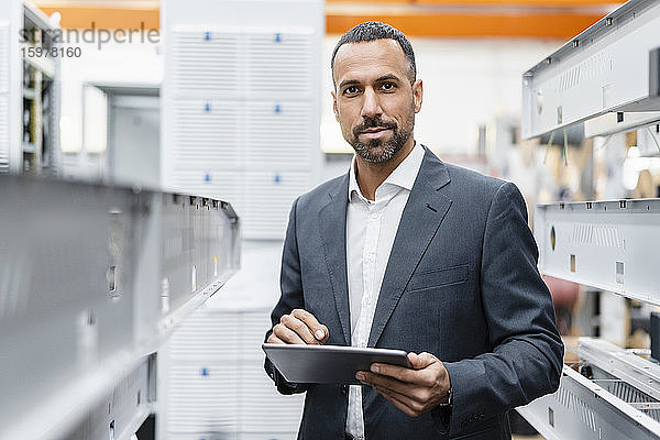 Porträt eines selbstbewussten Geschäftsmannes  der ein Tablet an Metallstangen in einer Fabrikhalle hält