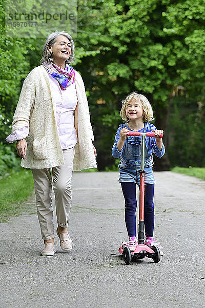 Glückliche ältere Frau  die mit ihrer Enkelin im Park spazieren geht und einen Motorroller fährt