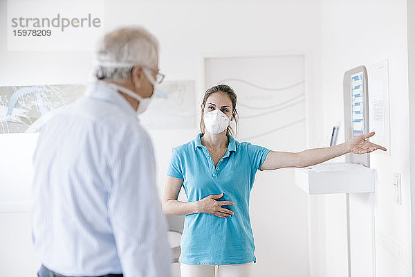 Ein älterer Mann trifft seine weibliche Physiotherapeutin an der Rezeption  die Gesichtsmasken trägt
