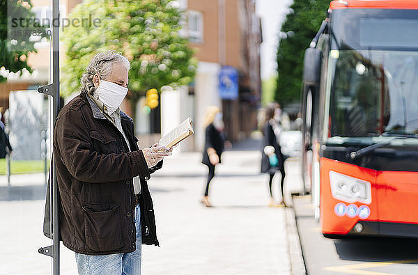 Älterer Mann mit Schutzmaske und Handschuhen liest ein Buch  während er an einer Bushaltestelle wartet  Spanien