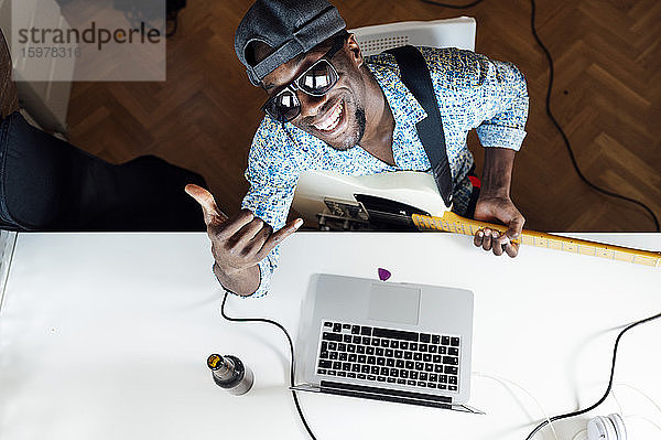 Porträt eines lächelnden jungen Mannes mit Shaka-Zeichen  der mit Gitarre und Laptop am Schreibtisch sitzt