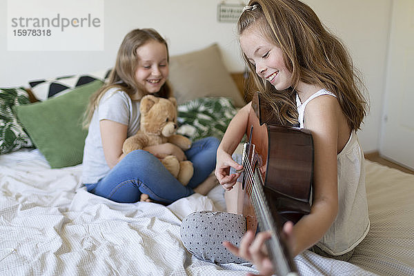 Lächelndes Mädchen sitzt auf dem Bett und spielt Gitarre  während ihre Schwestern zuhören