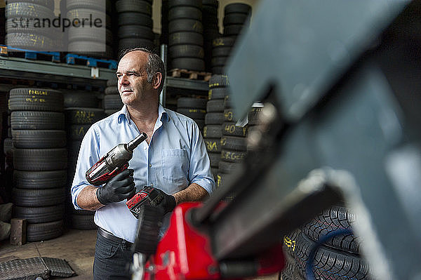 Ein älterer Unternehmer hält einen Schlagschrauber in der Hand  während er in einem Reifengeschäft wegschaut