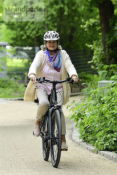 Lächelnde ältere Frau fährt mit dem Elektrofahrrad auf dem Fußweg im Park