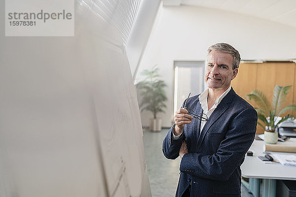 Porträt eines lächelnden männlichen Unternehmers  der eine Brille hält und an einem Whiteboard im Büro steht