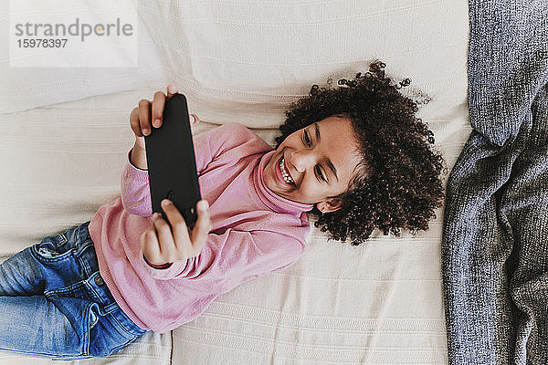 Porträt eines glücklichen kleinen Mädchens  das auf der Couch liegt und sein Smartphone benutzt