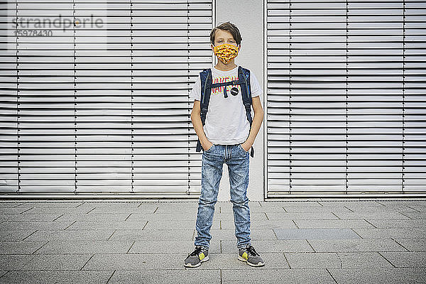 Junge mit Schultasche und Maske vor einem Gebäude stehend