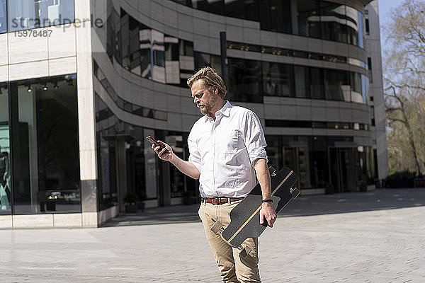 Geschäftsmann hält Skateboard und überprüft Smartphone in der Stadt