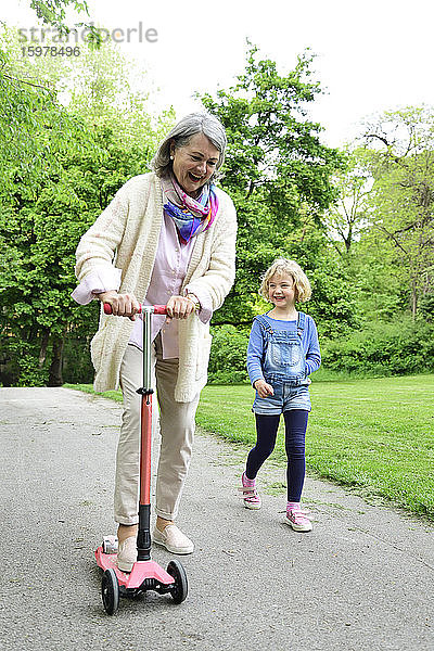 Fröhliche ältere Frau fährt mit dem Roller  während ihre Enkelin im Park spazieren geht
