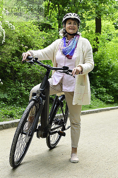 Nachdenkliche ältere Frau mit Elektrofahrrad auf dem Fußweg im Park stehend