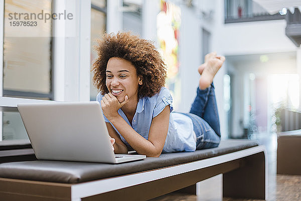 Junge Frau liegt auf einer Bank in einem modernen Büro und benutzt einen Laptop