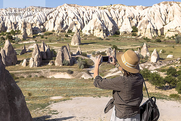 Seitenansicht einer jungen Frau  die mit ihrem Smartphone die felsige Landschaft im Dorf Goreme  Kappadokien  Türkei  fotografiert