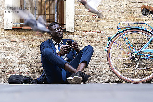 Junger Geschäftsmann mit Smartphone auf dem Boden sitzend in der Stadt