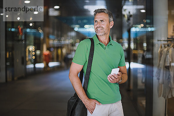 Lächelnder reifer Mann  der wegschaut  während er sein Smartphone im Einkaufszentrum benutzt