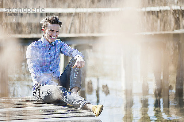 Gut aussehender erwachsener Mann  der wegschaut  während er auf einem Steg über einem See sitzt