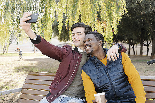 Zwei glückliche junge Männer sitzen auf einer Parkbank und machen ein Selfie