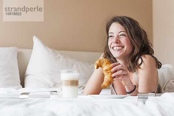 Glückliche junge Frau mit Croissant in der Hand beim Frühstück im Bett