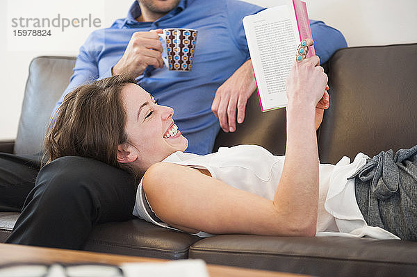 Fröhliche Frau liest ein Buch  während sie auf dem Schoß eines Mannes auf der Couch zu Hause liegt