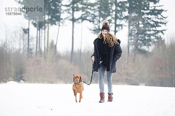 Lächelnde junge Frau  die mit ihrem Hund im Schnee vor Bäumen im Wald spazieren geht