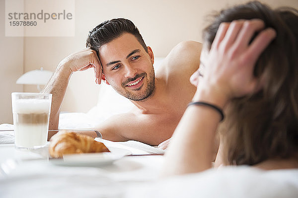 Glücklicher junger Mann  der eine Frau ansieht  während er beim Frühstück zu Hause im Bett liegt