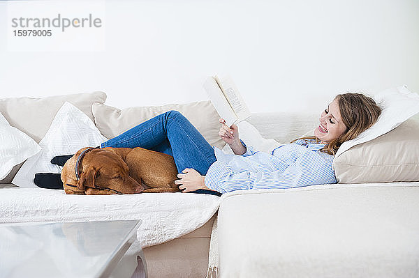 Lächelnde junge Frau liegt mit Buch und Hund auf dem Sofa im Wohnzimmer zu Hause